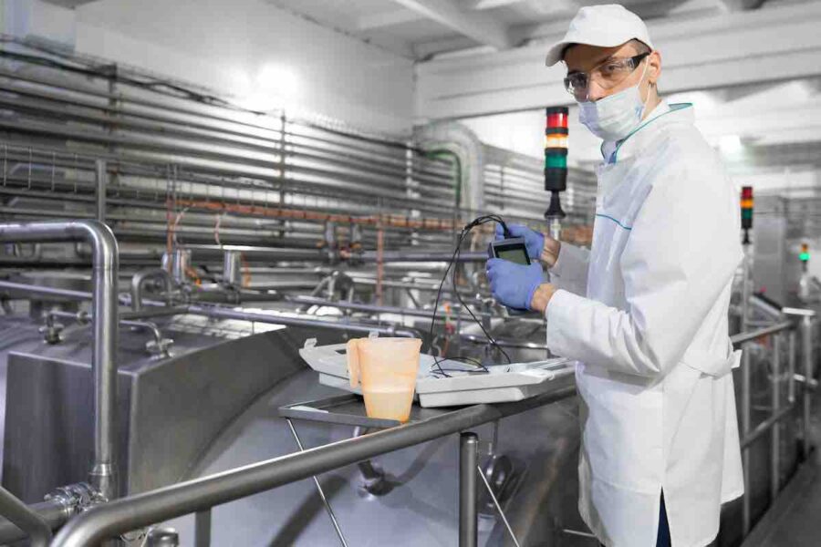 man in a white coat, gloves and a mask stands near the production line and holds a tester with wires at the dairy plant. The inspector carries out control at the cheese factory